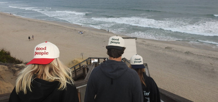 Three people wearing 'Good people' hats looking at the ocean from a beach.