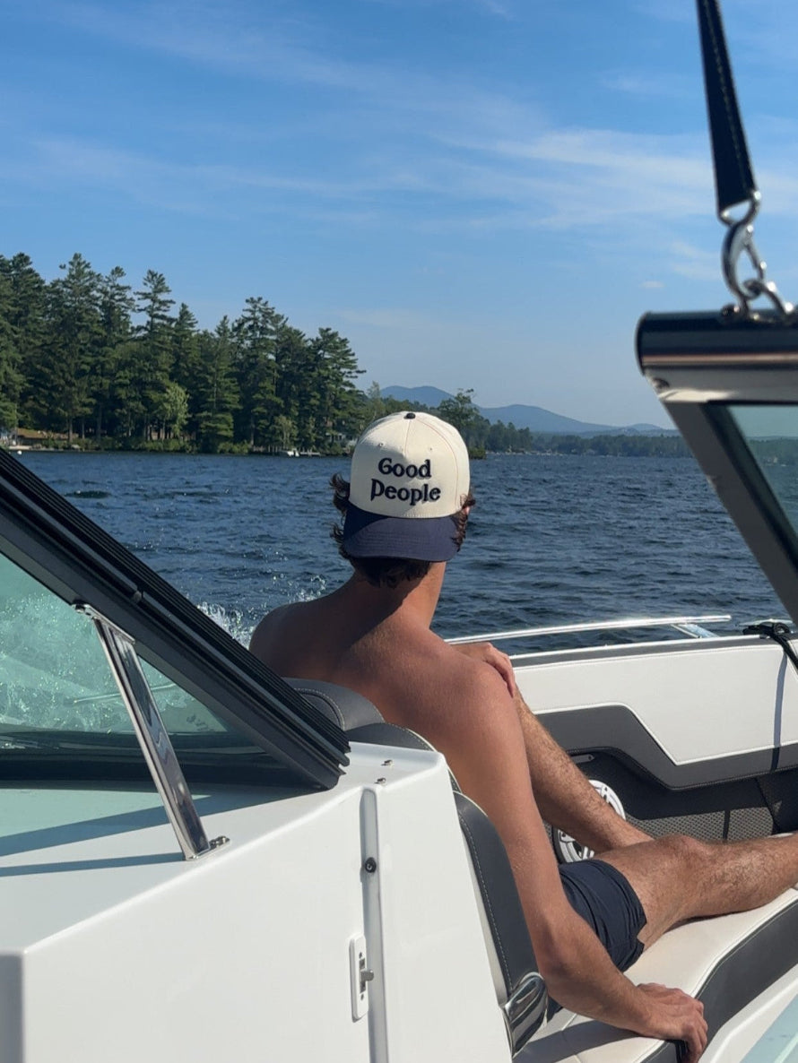 Person on a boat wearing a cap with 'Good People' text, looking at a scenic lake with trees and mountains.