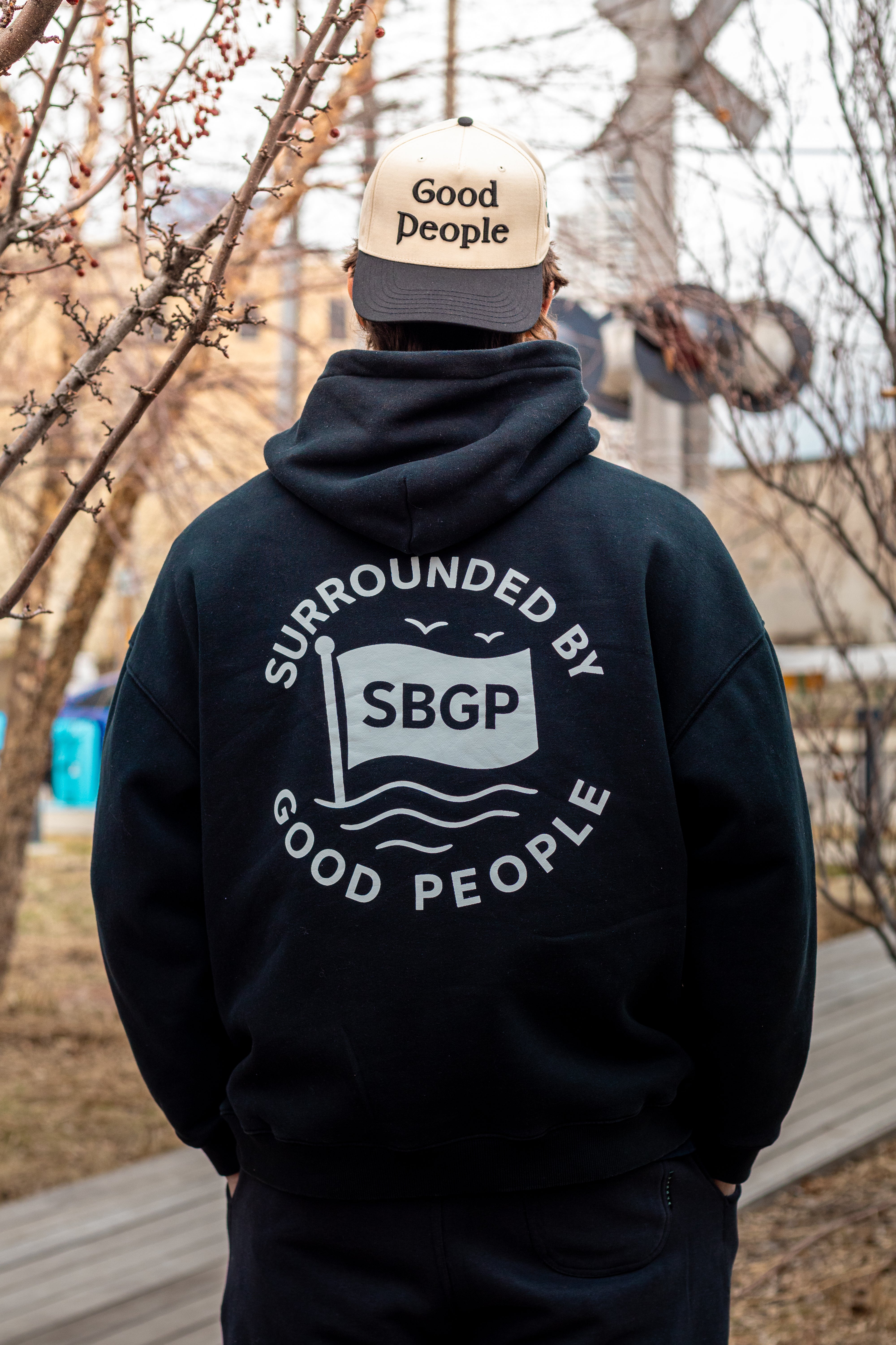 Person wearing a black hoodie with 'Surrounded by Good People' text and a cap with 'Good People' text, standing outdoors.